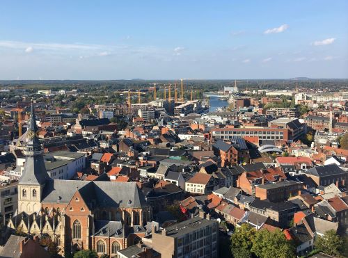Een panoramisch uitzicht over de stad Hasselt met kerken, daken van gebouwen en in de verte bouwkranen, onder een blauwe lucht