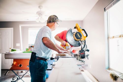 Een man met een pet en veiligheidsbril bedient een verstekzaag terwijl hij aan een houten plank werkt in een licht gevulde kamer tijdens een woningrenovatie.