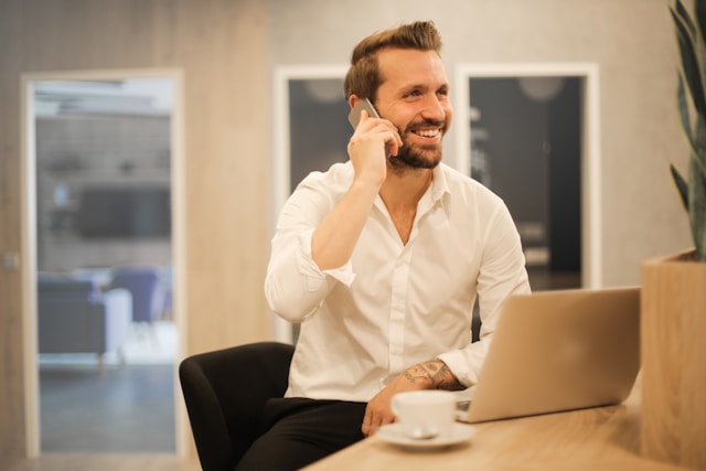 Een lachende man in een wit overhemd die aan een bureau zit met een laptop en een kop koffie, terwijl hij telefoneert.