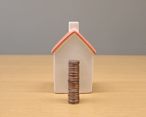 Een stapel munten voor een klein huisvormig spaarpot op een houten tafel.