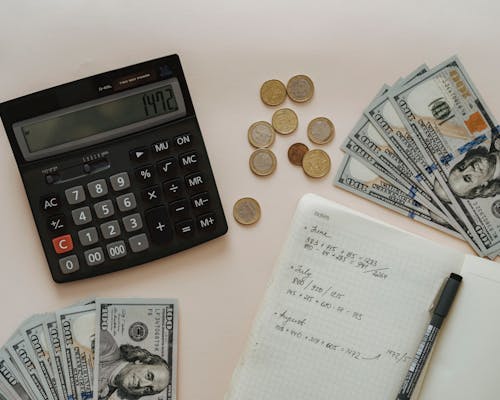 Een rekenmachine, munten, dollarbiljetten en een notitieboekje met berekeningen op een tafel.